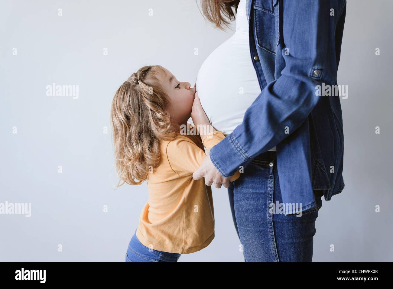 Cute blond girl kissing pregnant mother's belly by wall Stock Photo