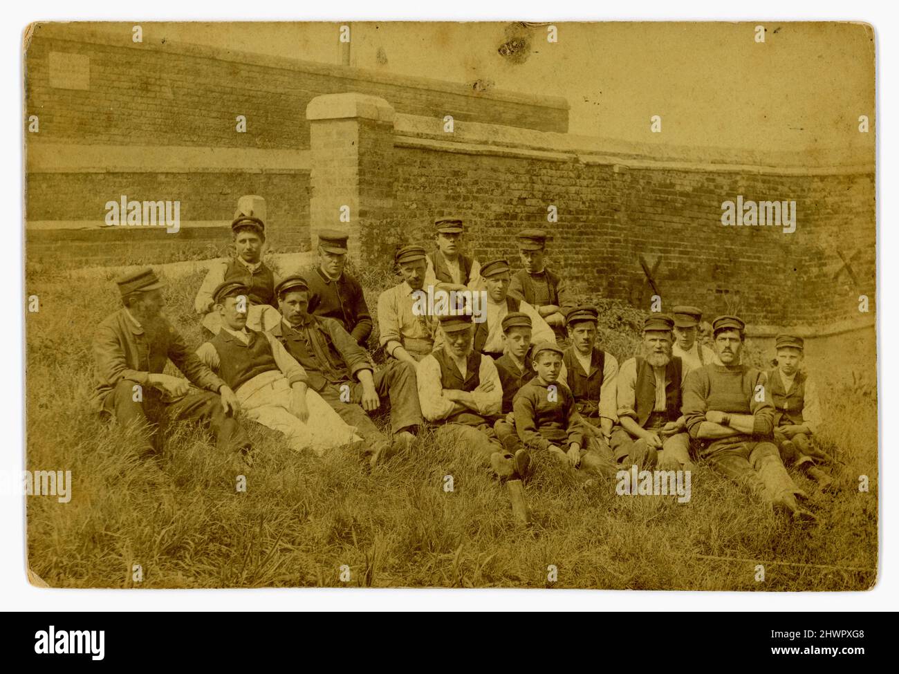 Original sepia-toned Victorian era cabinet card relaxed outdoor portrait of large group of working class men and boys (apprenticed maybe) sitting near an ancient harbour wall near a slipway. They are all wearing peaked mariners caps, and some are wearing hand knitted fishermen's sweaters, another an apron, and are probably in the fishing industry - a coastal seafaring fishing community in the U.K. Dated June 1892. Stock Photo