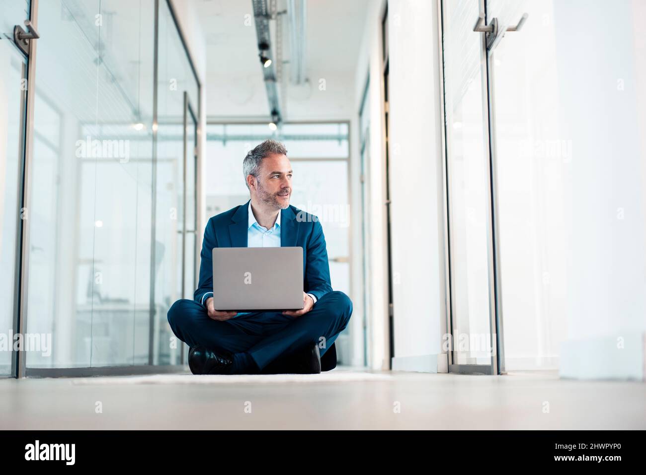Thoughtful businessman sitting cross-legged with laptop in office corridor Stock Photo
