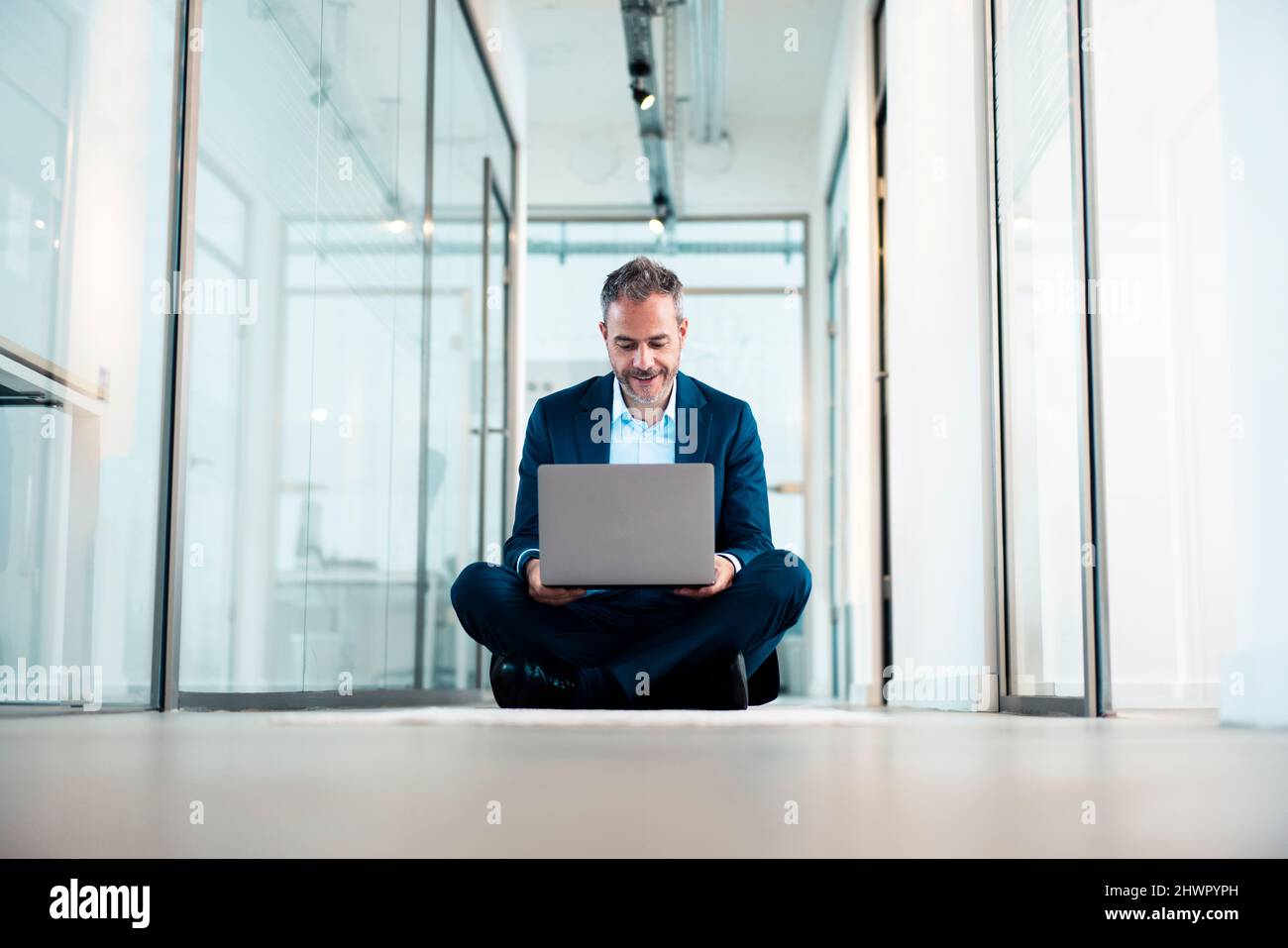Businessman sitting cross-legged using laptop in office corridor Stock Photo