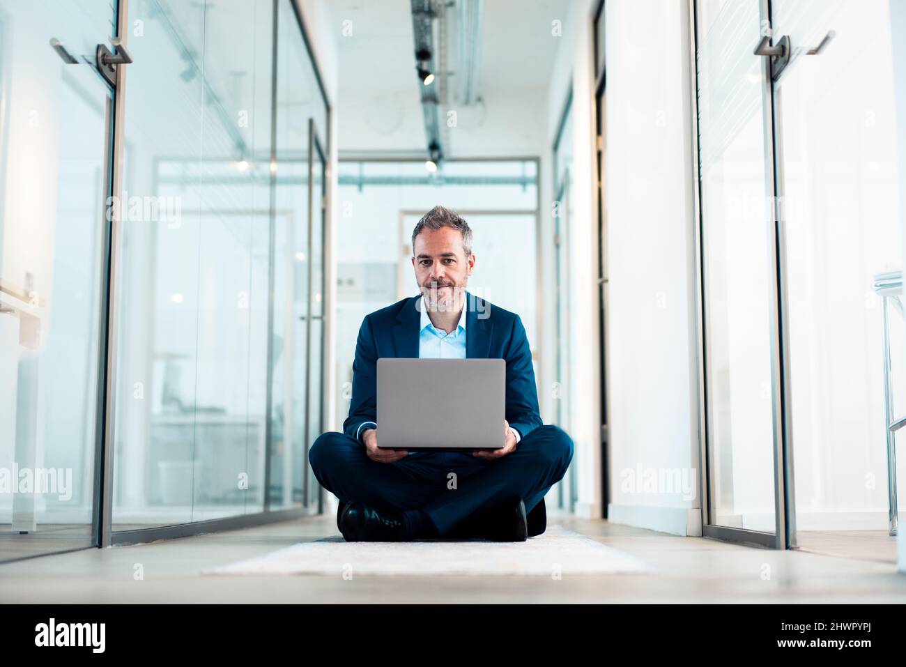 Confident businessman sitting cross-legged with laptop in office corridor Stock Photo