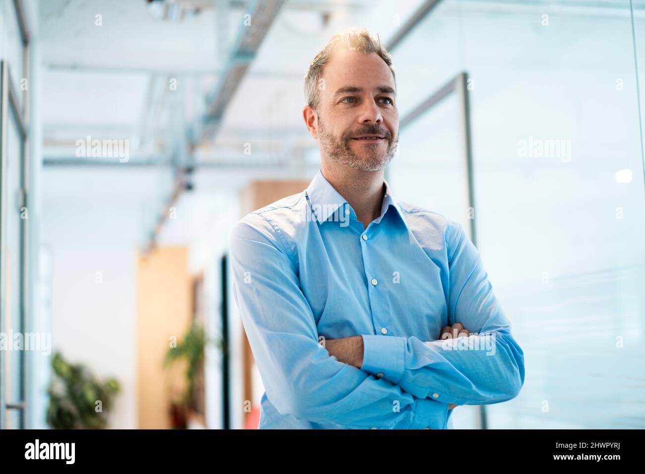 Thoughtful businessman with arms crossed standing in corridor at ...
