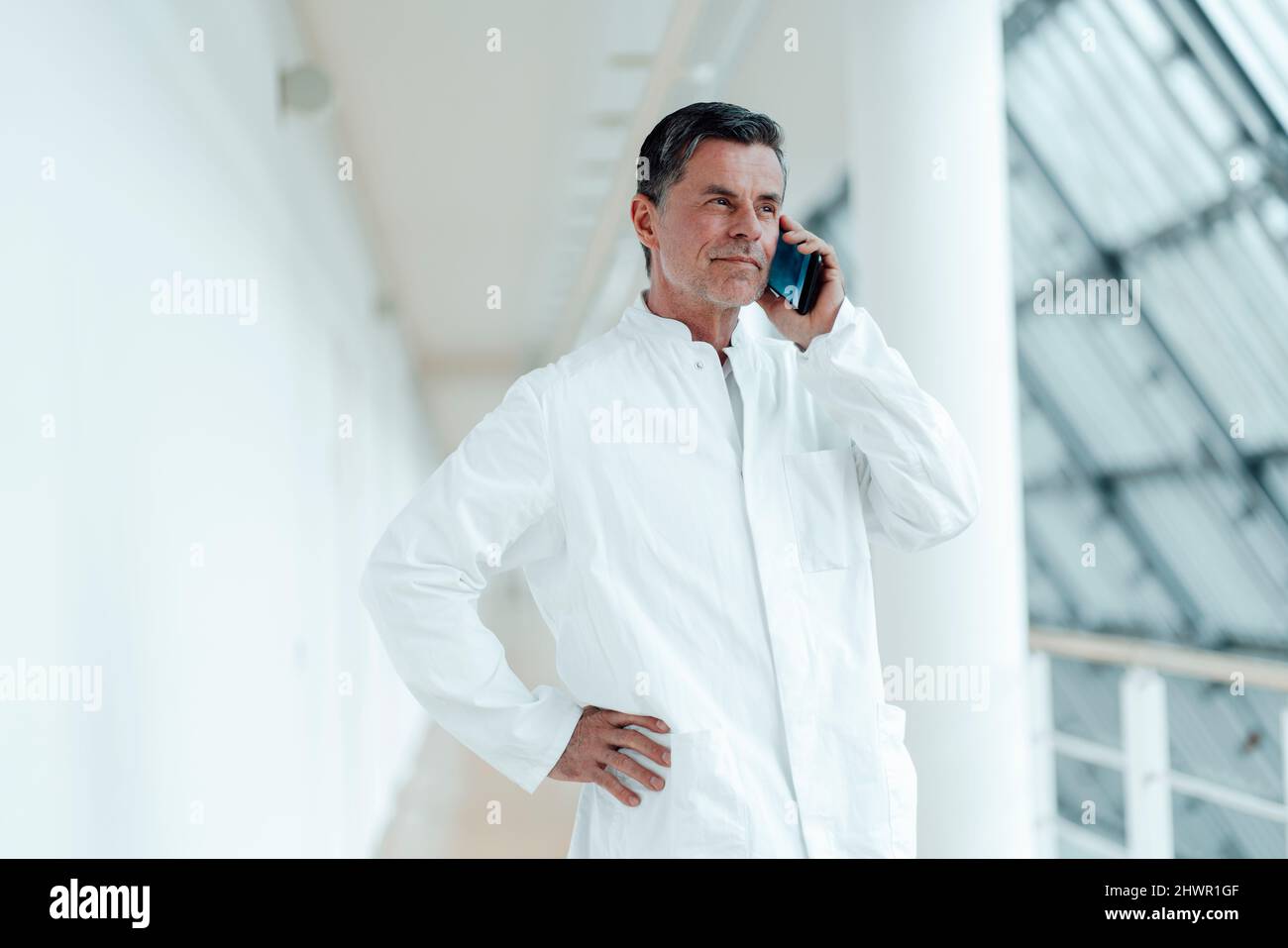 Scientist with hand on hip talking on smart phone by column at medical clinic Stock Photo