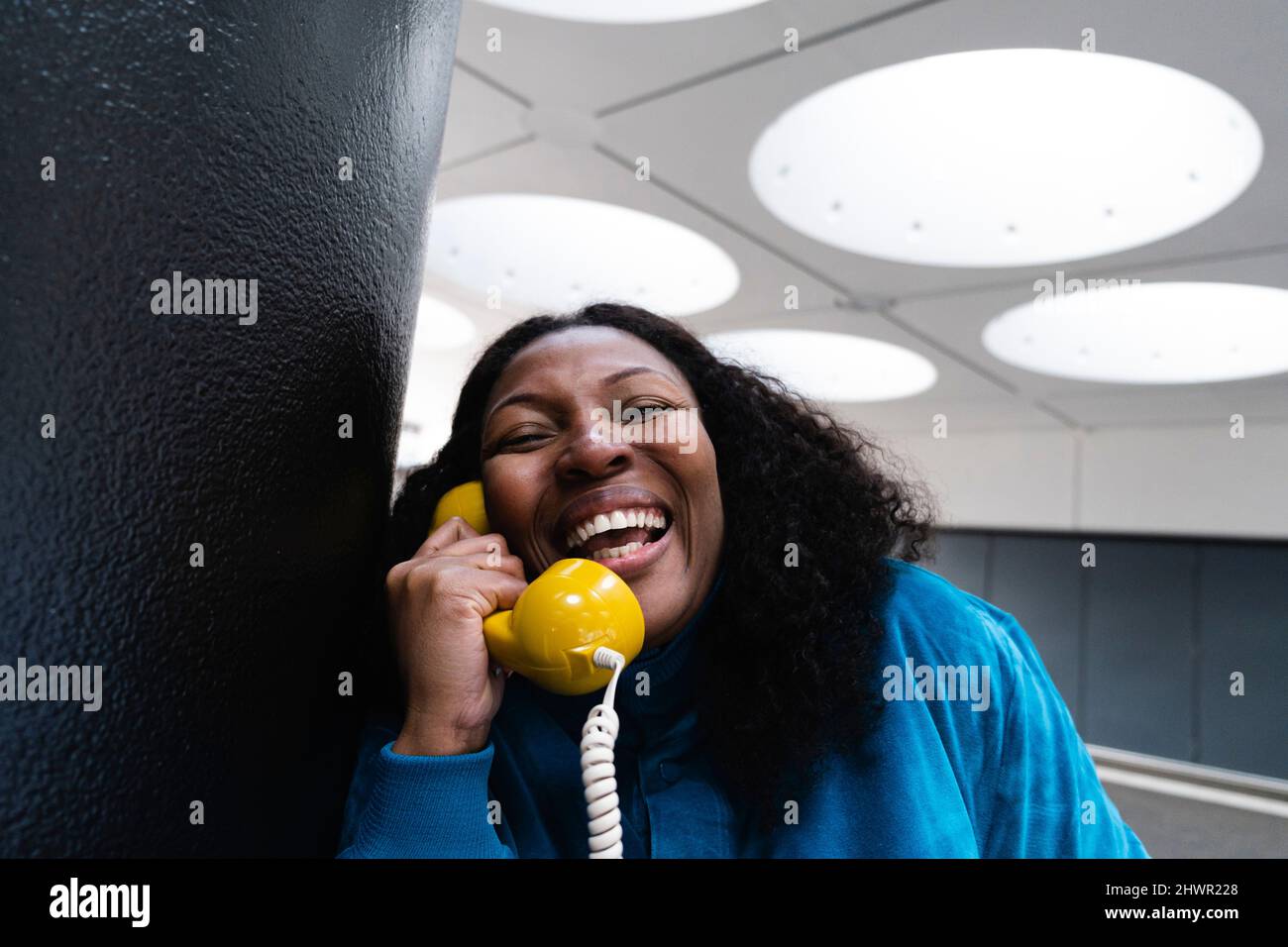 Happy woman listening through old-fashioned yellow telephone receiver leaning on column Stock Photo