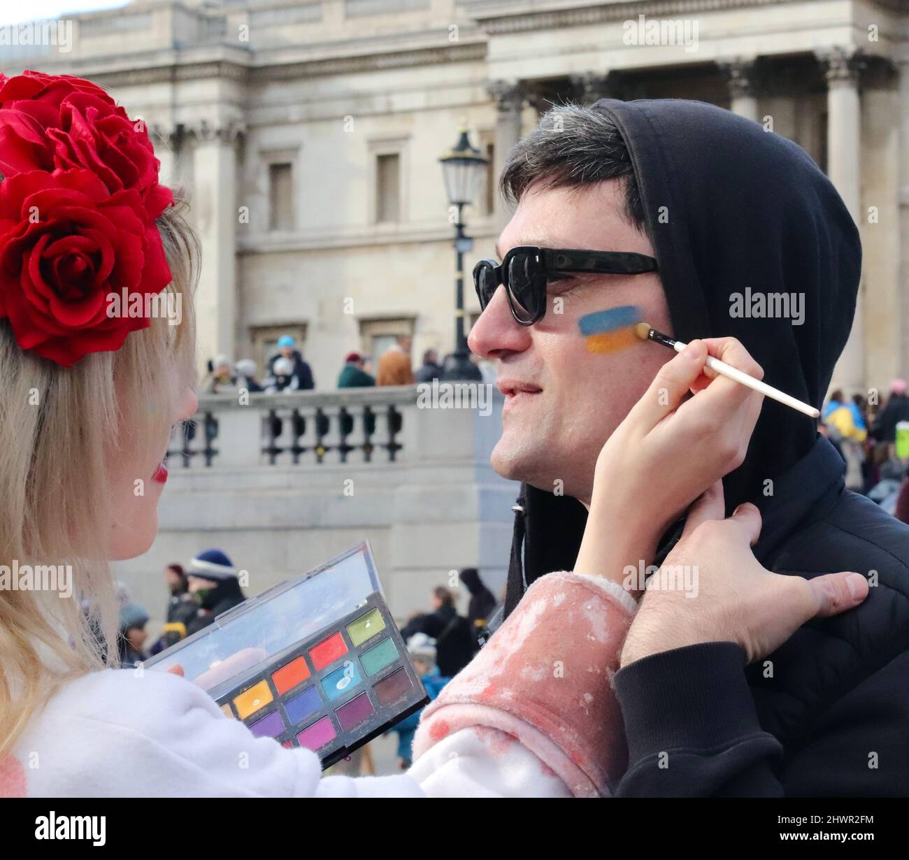 Stand with Ukraine rally in Trafalgar Square, London 2022. Lady paints Ukrainian flag on mans face to show solidarity with Ukraine. Stock Photo