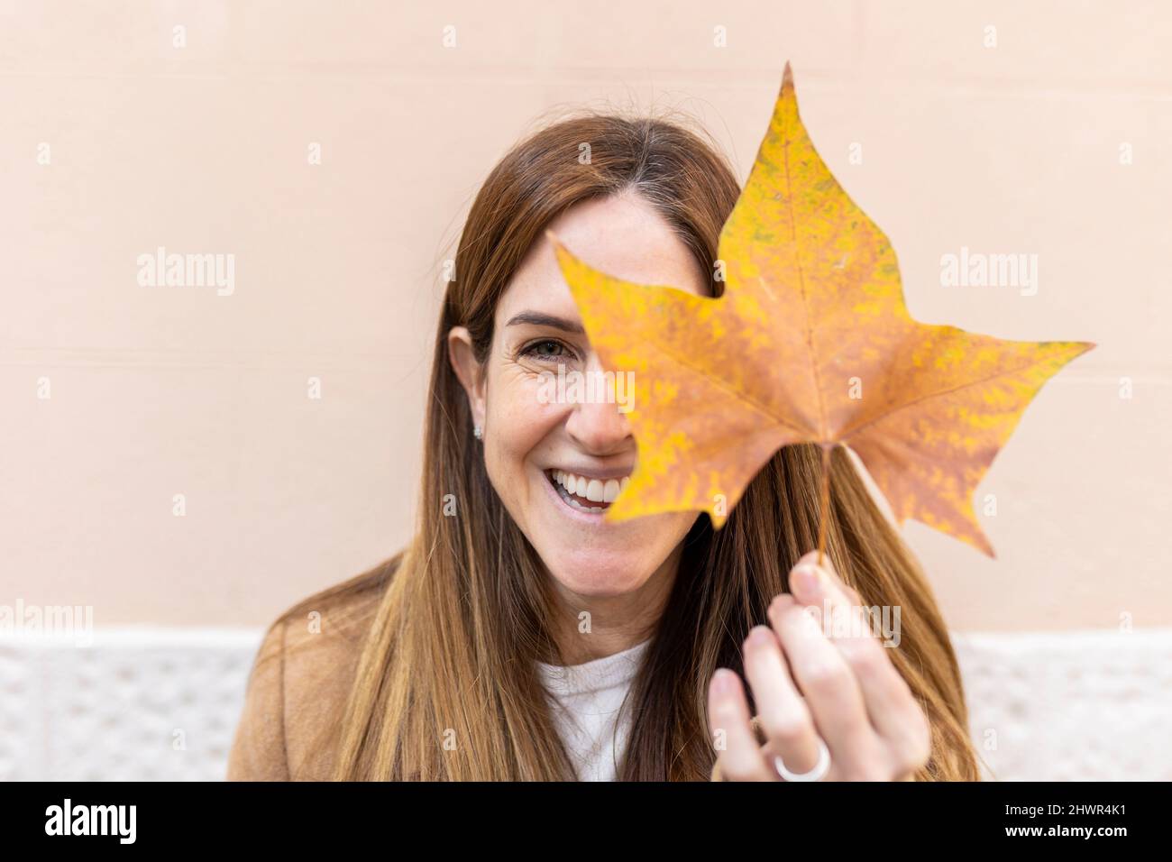 Cheerful woman covering eye with autumn leaf in front of wall Stock ...
