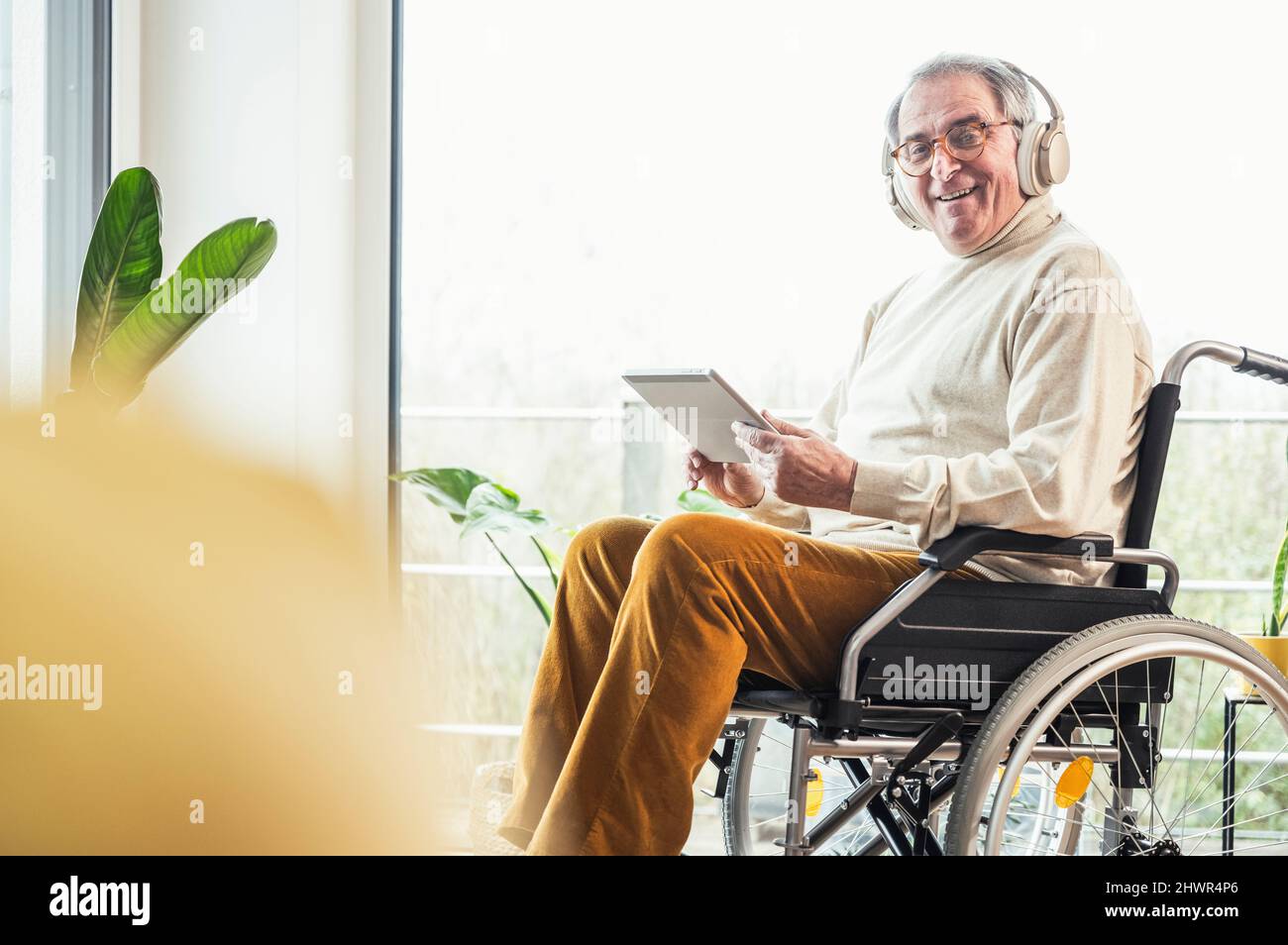 Smiling senior disabled man with tablet PC sitting on wheelchair at home Stock Photo