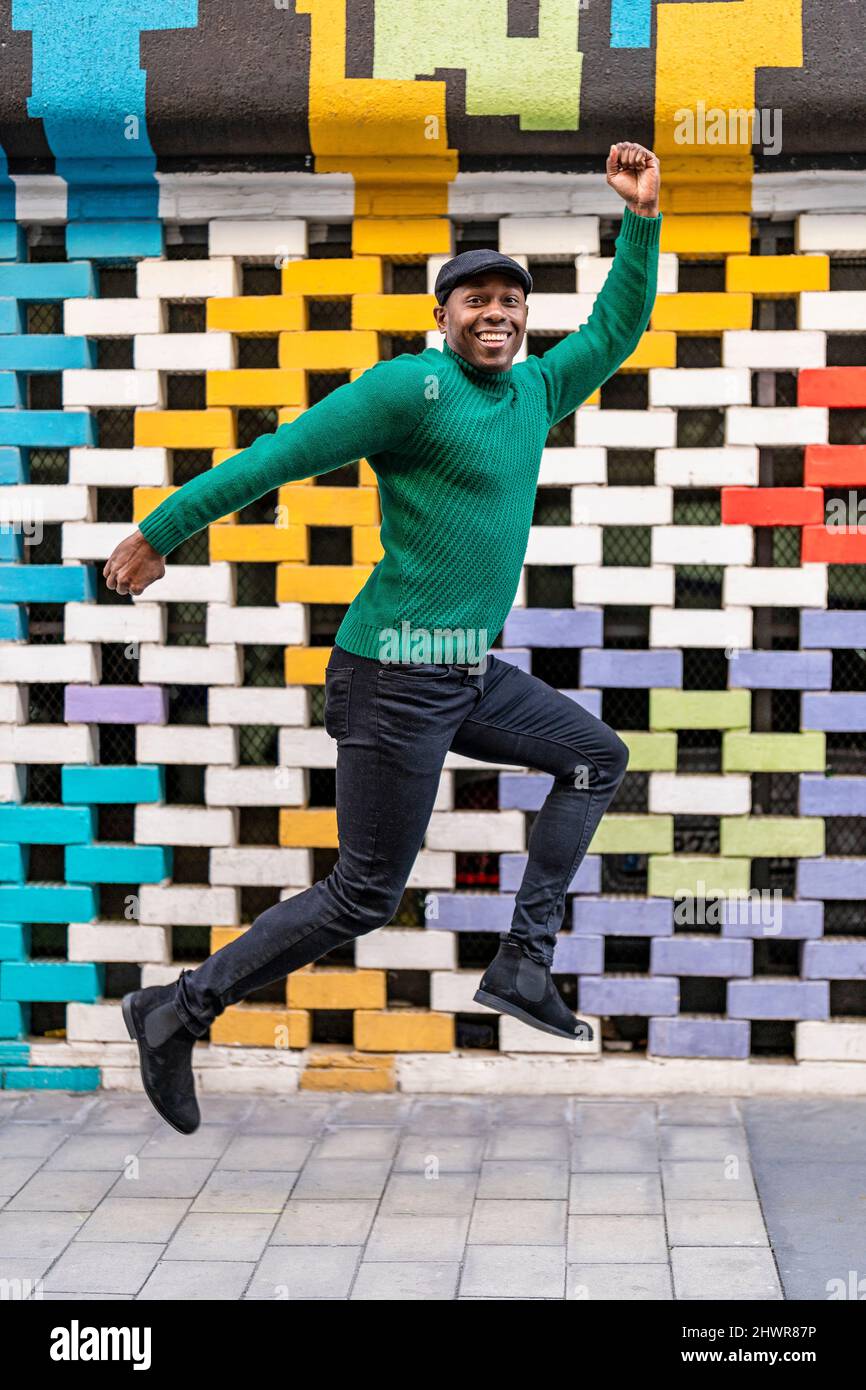 Happy man wearing flat cap jumping in front of multi colored wall Stock Photo