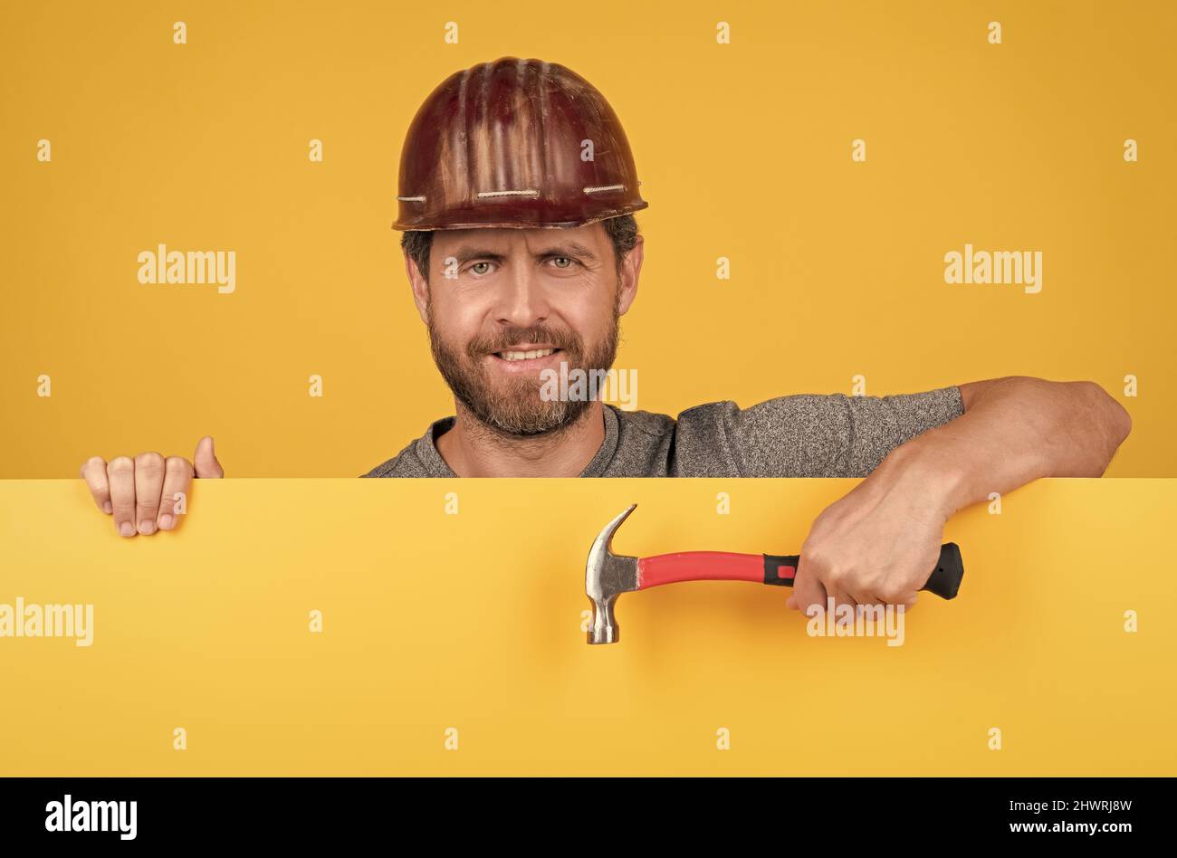 happy mature man laborer in helmet behind yellow paper for copy space hold hammer, happy workers day Stock Photo