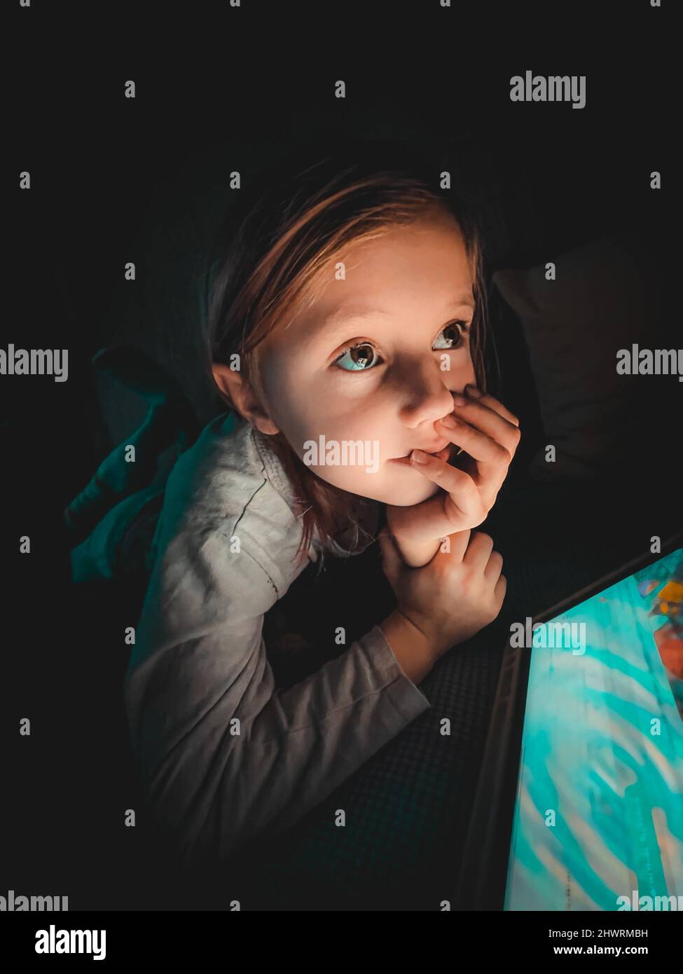 Little caucasian girl looking at a tablet in the evening in the dark on the bed,lifestyle toned photography at home, selective focus and softfocus Stock Photo