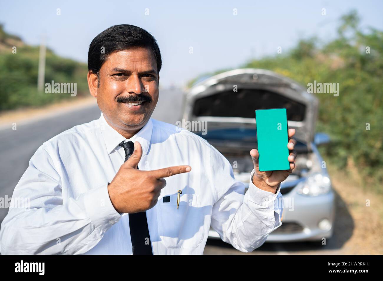 Happy businessman showing green screen mobile phone in front of broken car by looking at camera - concept of online mobile car repair service booking Stock Photo