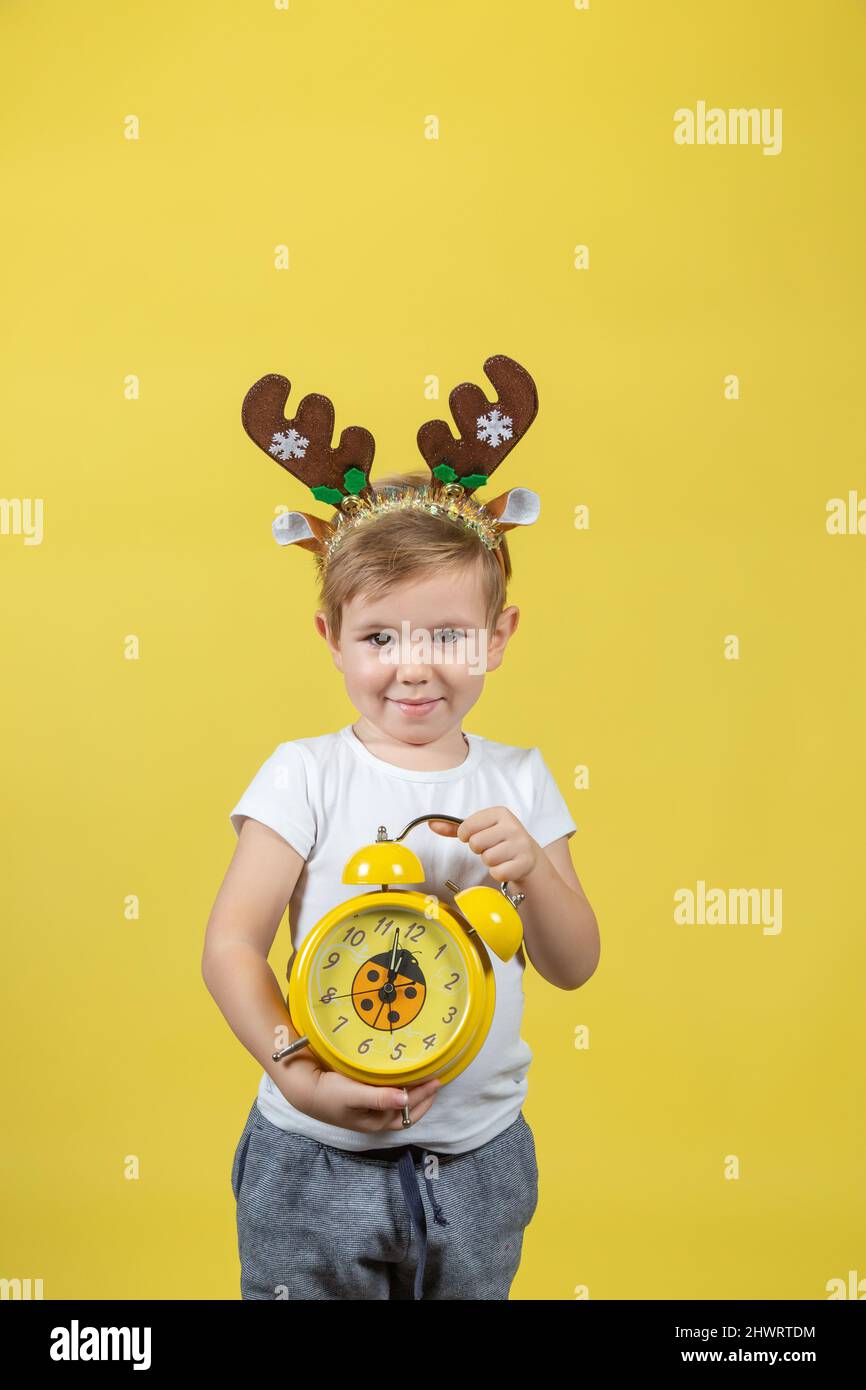 A smiling little boy in in christmas reindeer costume with alarm clock in his hands. A place for your text. Studio shot on a yellow background. The co Stock Photo