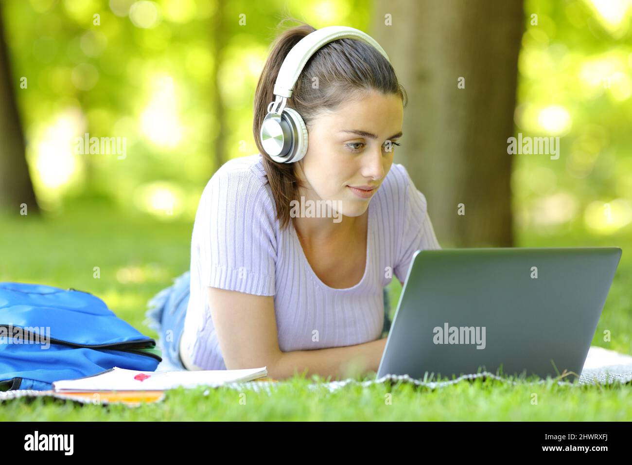 Woman e-learning online using laptop and headphones lying on grass in a park in a campus Stock Photo