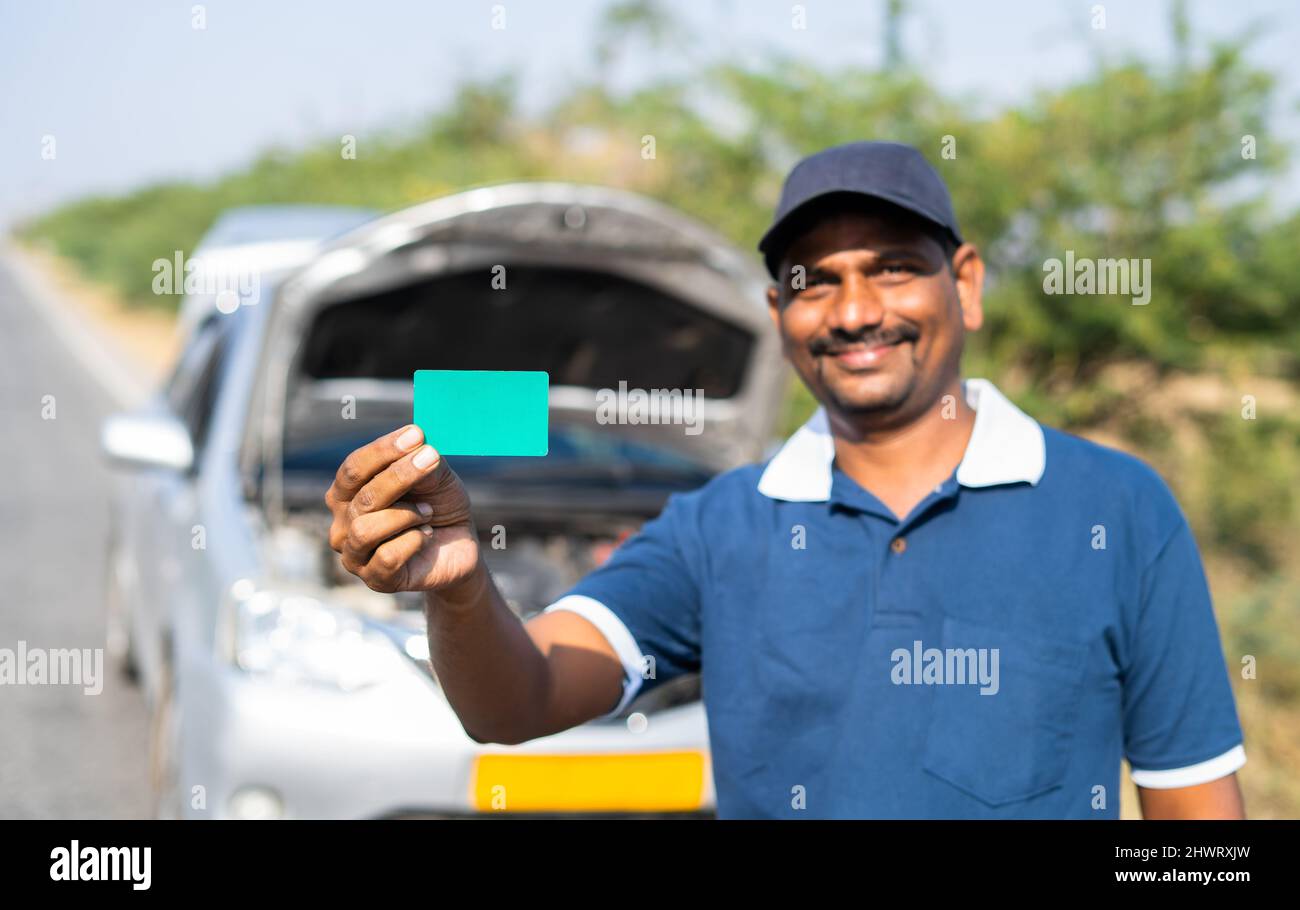 Smiling car mechanic showing green card by looking at camera in front of broken car - concept of promotion for mobile repair service. Stock Photo