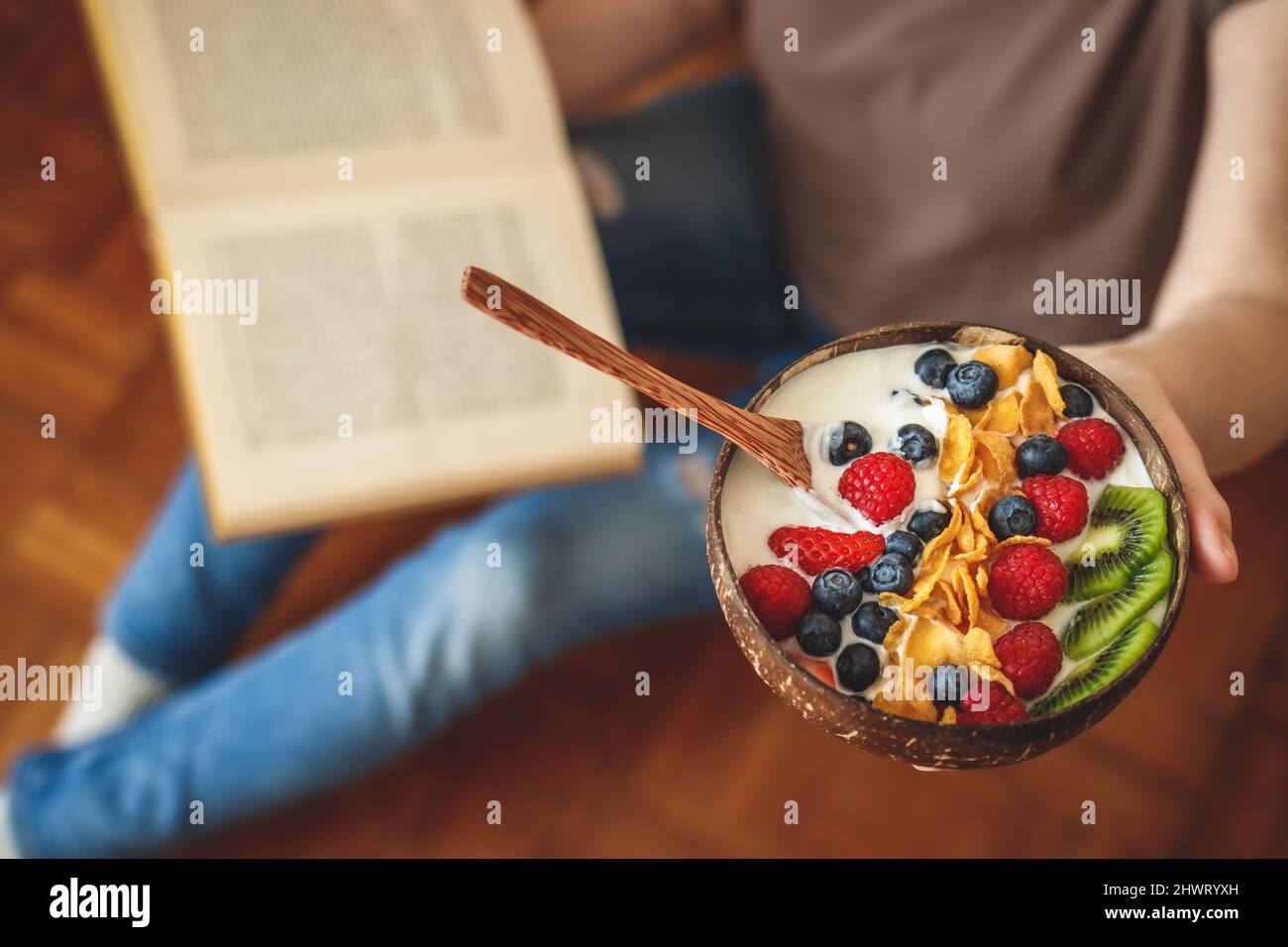 Yogurt, fruit and corn flakes in coconut bowl. Woman reading book during breakfast at home. Healthy eating and lifestyle Stock Photo