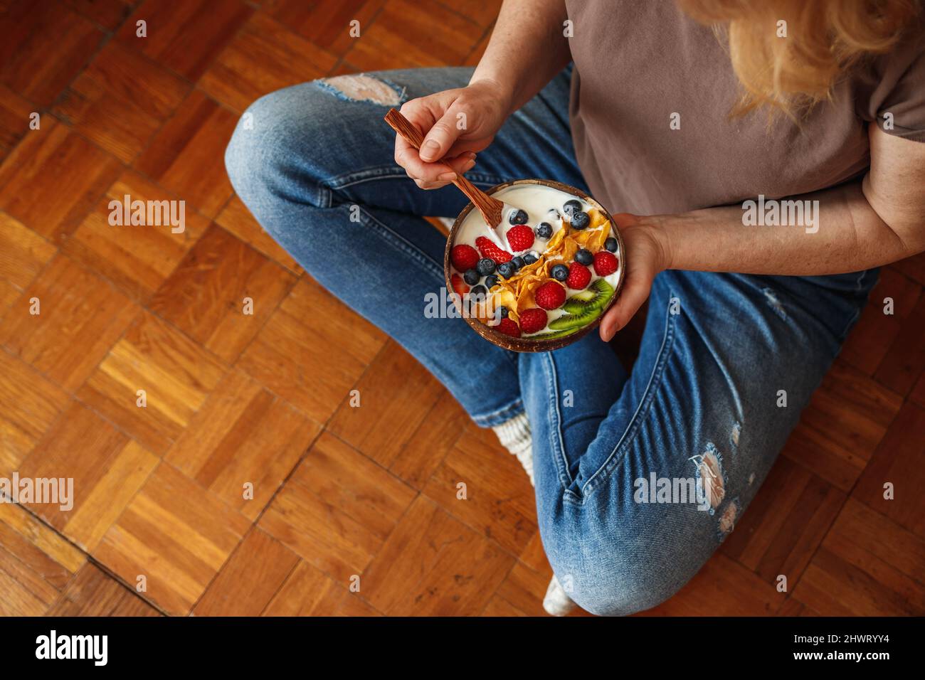 Healthy breakfast at home. Woman sitting on floor and eating yoghurt with fruit and corn flakes. Vegetarian food Stock Photo