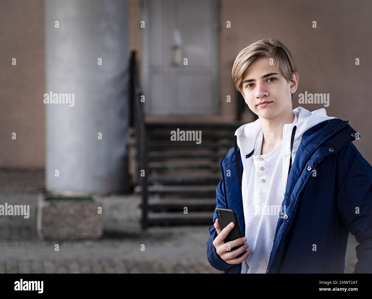 Teenager boy in a blue jacket and a white T-shirt holds a cell phone in his hands, looks at the camera and smiles. Stock Photo