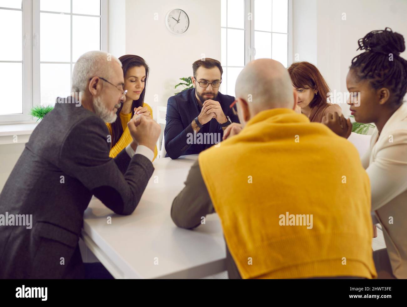 Group of different men and women sitting around table and praying to