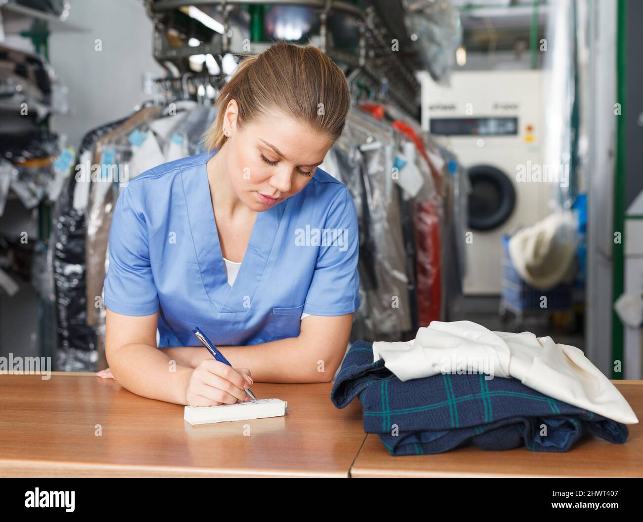 Focused girl worker of laundry writing receipt while taking clothes for dry cleaning Stock Photo