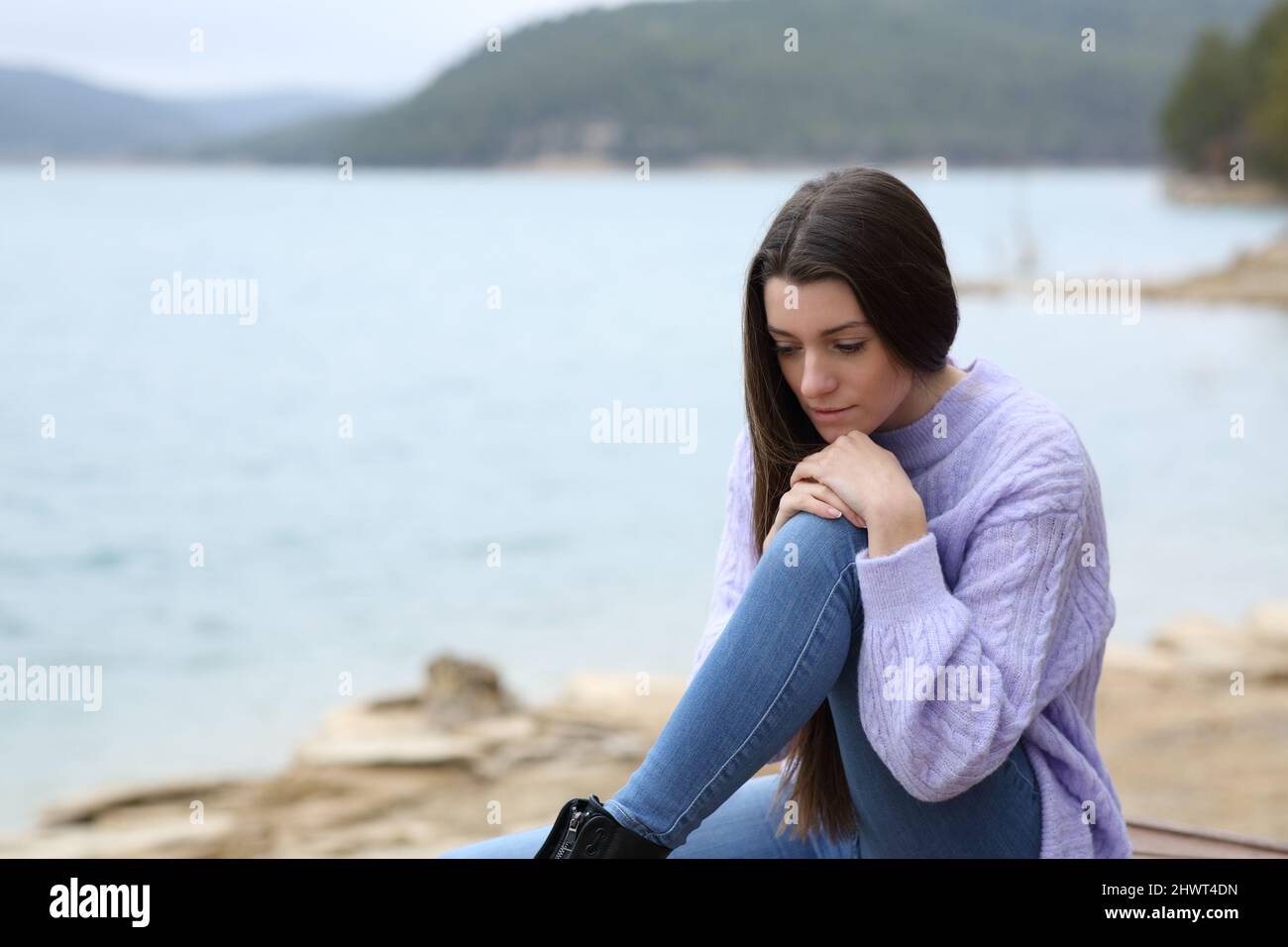 Sad pensive teen looking down sitting in a lake a cloudy day Stock Photo