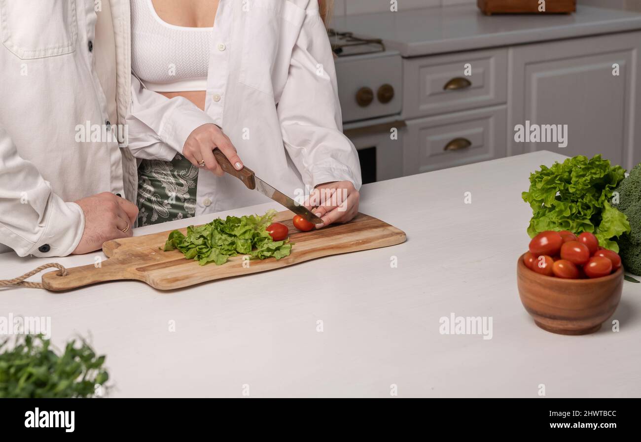 Couple cooking at home. Woman cutting vegetables for salad in modern white scandi kitchen. Healthy food concept. High quality photo Stock Photo