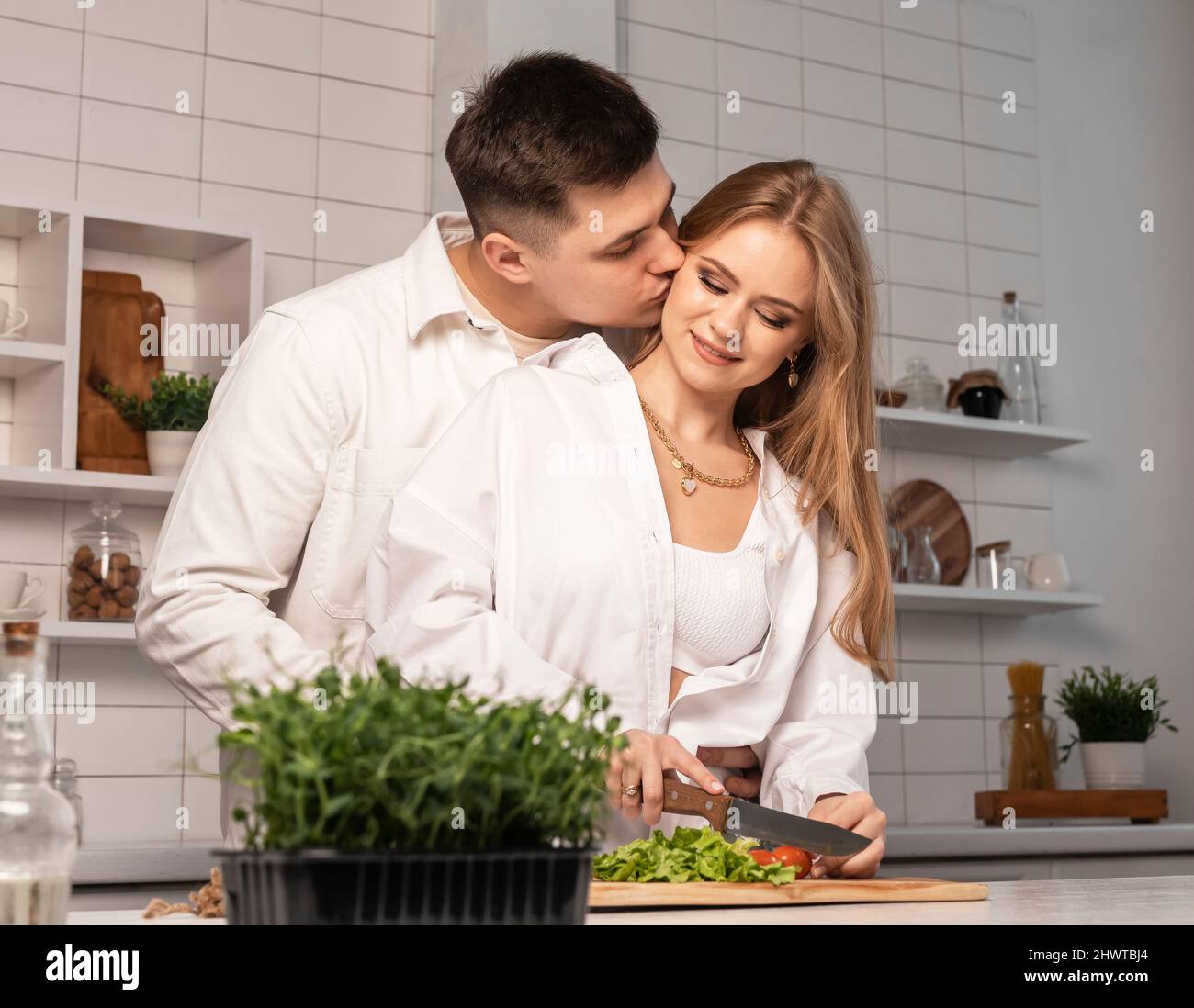 Romantic couple cooking together. Handsome man kissing his wife or girlfriend while woman cutting tomatoes for vegetables salad. High quality photo Stock Photo