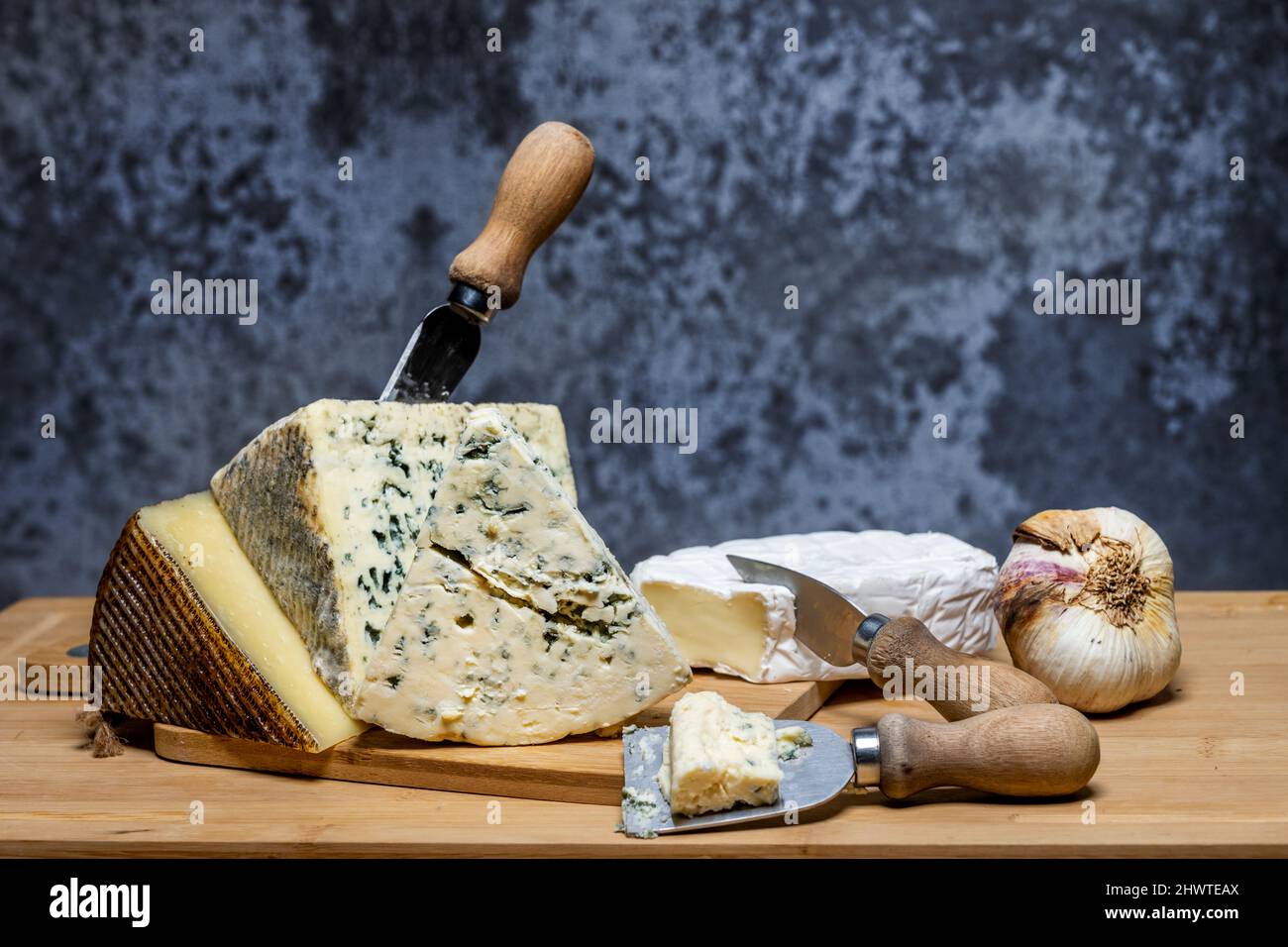 Still life with tacos of delicious and fragrant blue cheeses and brie cheese, a cured Manchego cheese and a head of garlic with small cheese knives an Stock Photo