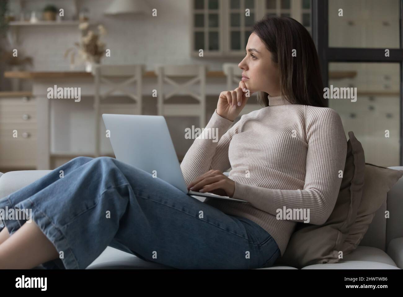 Thoughtful woman sit on couch with laptop staring away Stock Photo