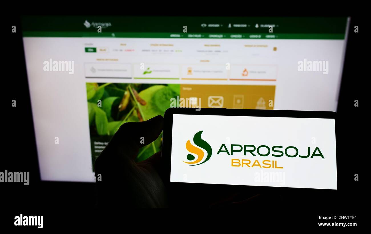 Person holding mobile phone with logo of Brazilian agriculture company Aprosoja on screen in front of business web page. Focus on phone display. Stock Photo
