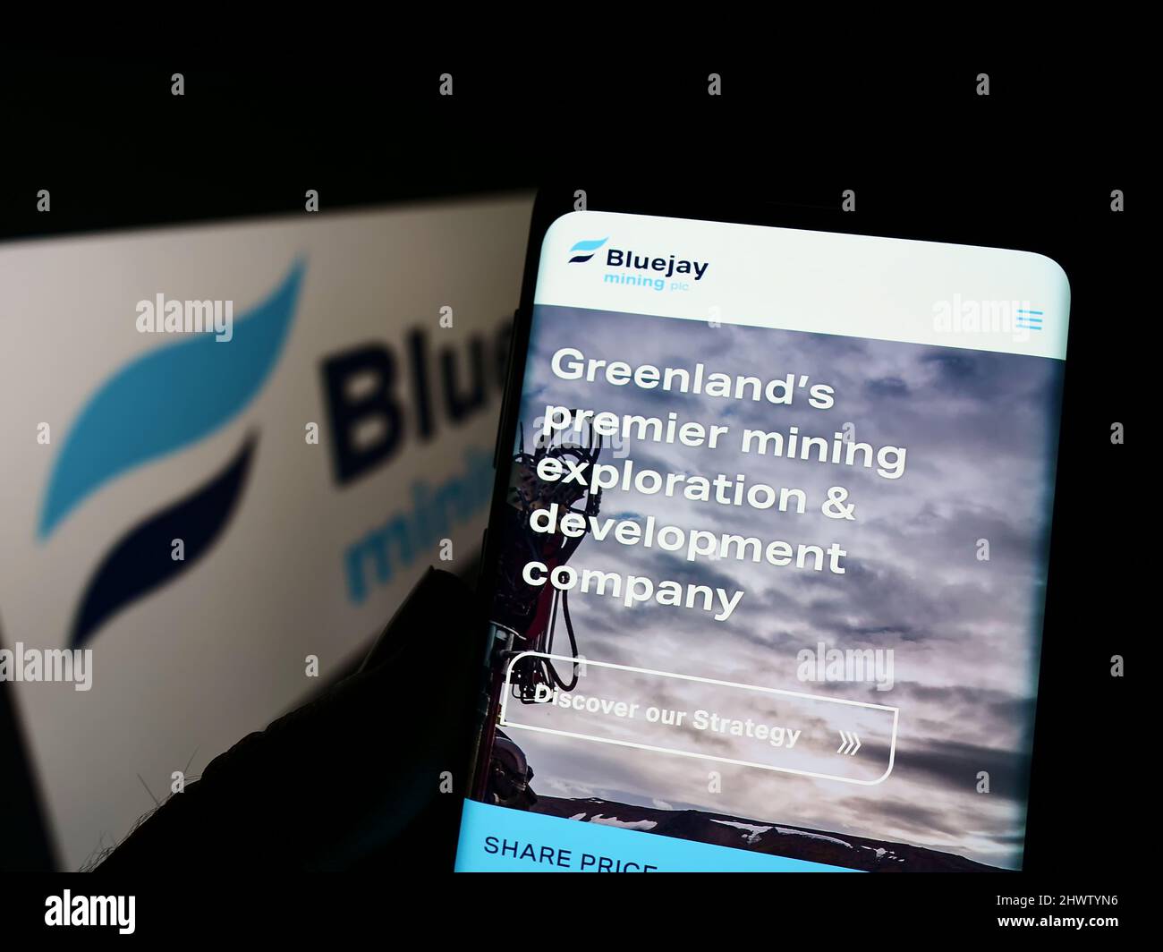 Person holding cellphone with website of British company Bluejay Mining plc on screen in front of logo. Focus on center of phone display. Stock Photo