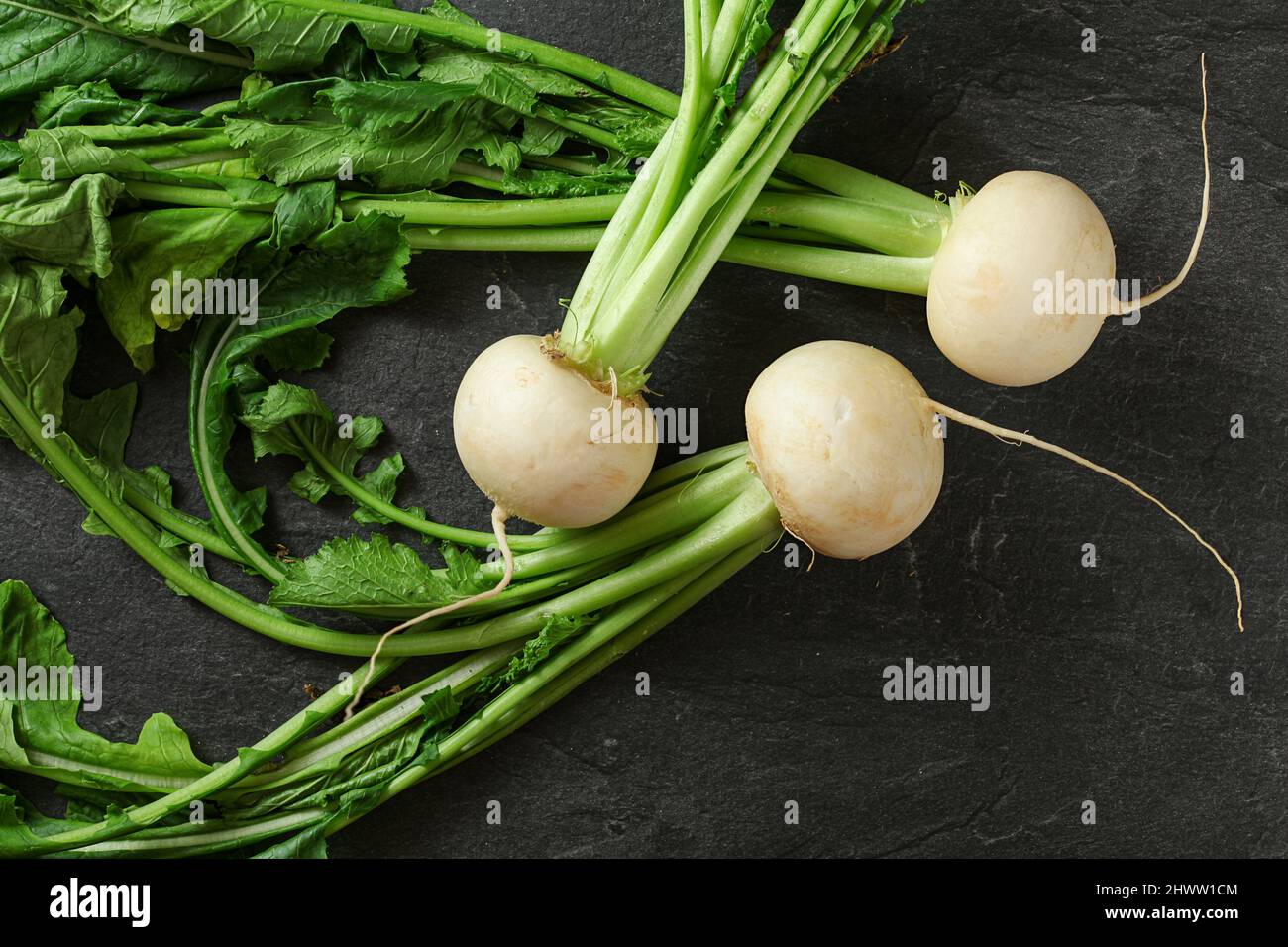 Three white radish bulbs with green leaves on black slate like board, view from above Stock Photo