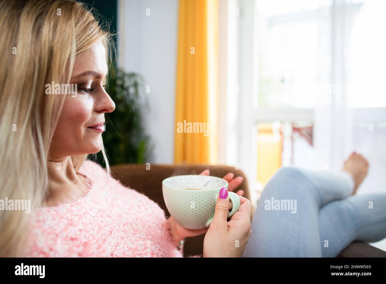 A blonde woman holds a cup of coffee while sitting in a chair. Stock Photo