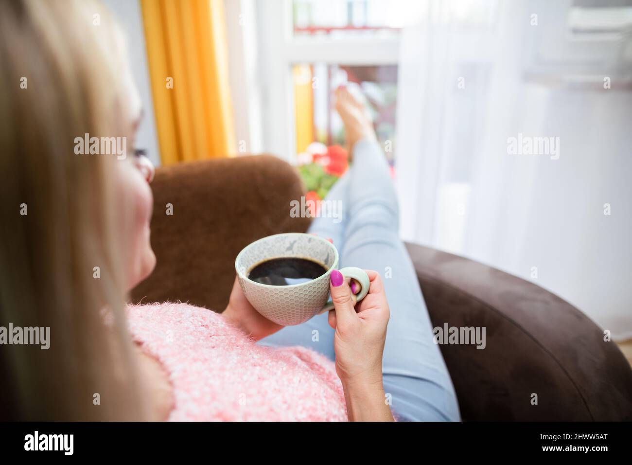 A blonde woman holds a cup of coffee while sitting in a chair. Stock Photo