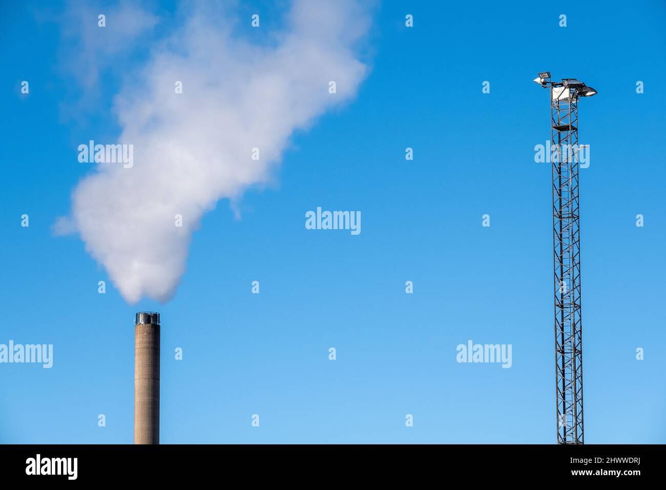 Helsinki / Finland - FEBRUARY 26, 2022: Tall light mast and a smokestack with exhaust gas against a bright blue sky Stock Photo