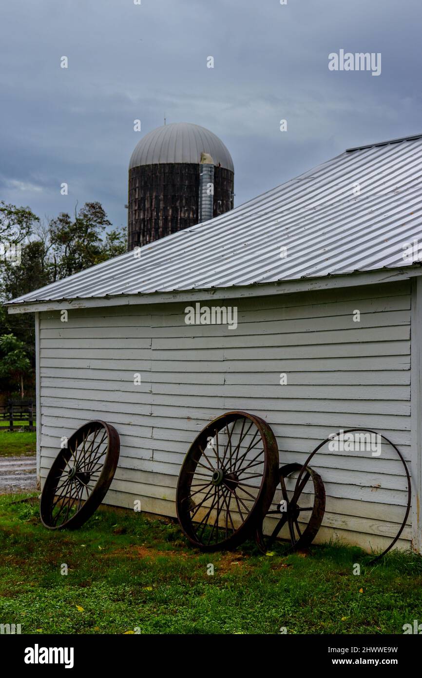 Old, rusty wagon wheels, lean against a white farm building with a silo in the background. Stock Photo