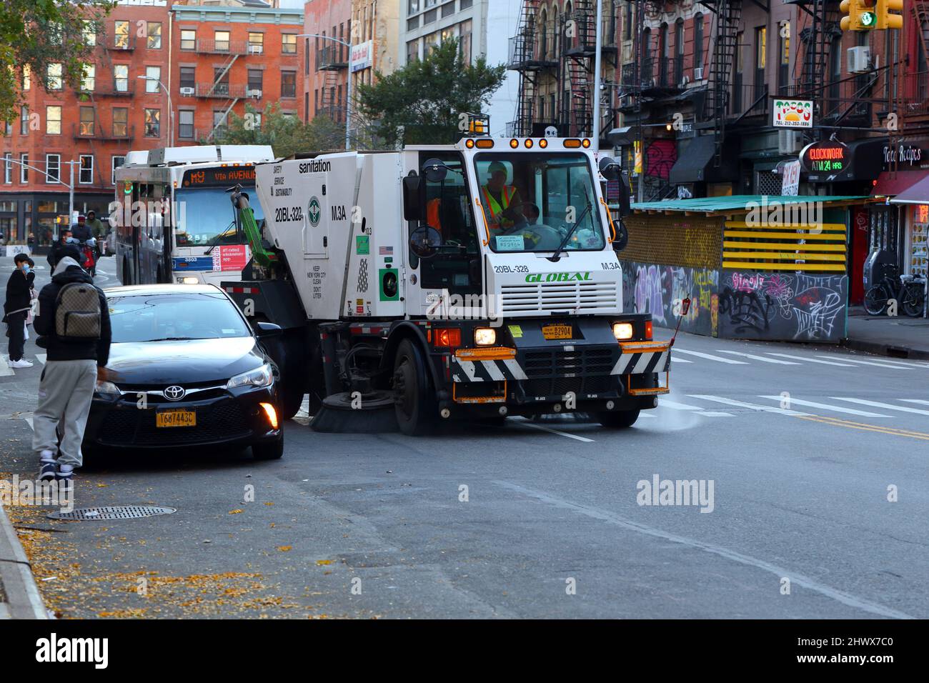 A NYC Sanitation street sweeper maneuvering around a ride-hail 'black car' standing away from the curb, blocking Essex St in the Lower East Side Stock Photo