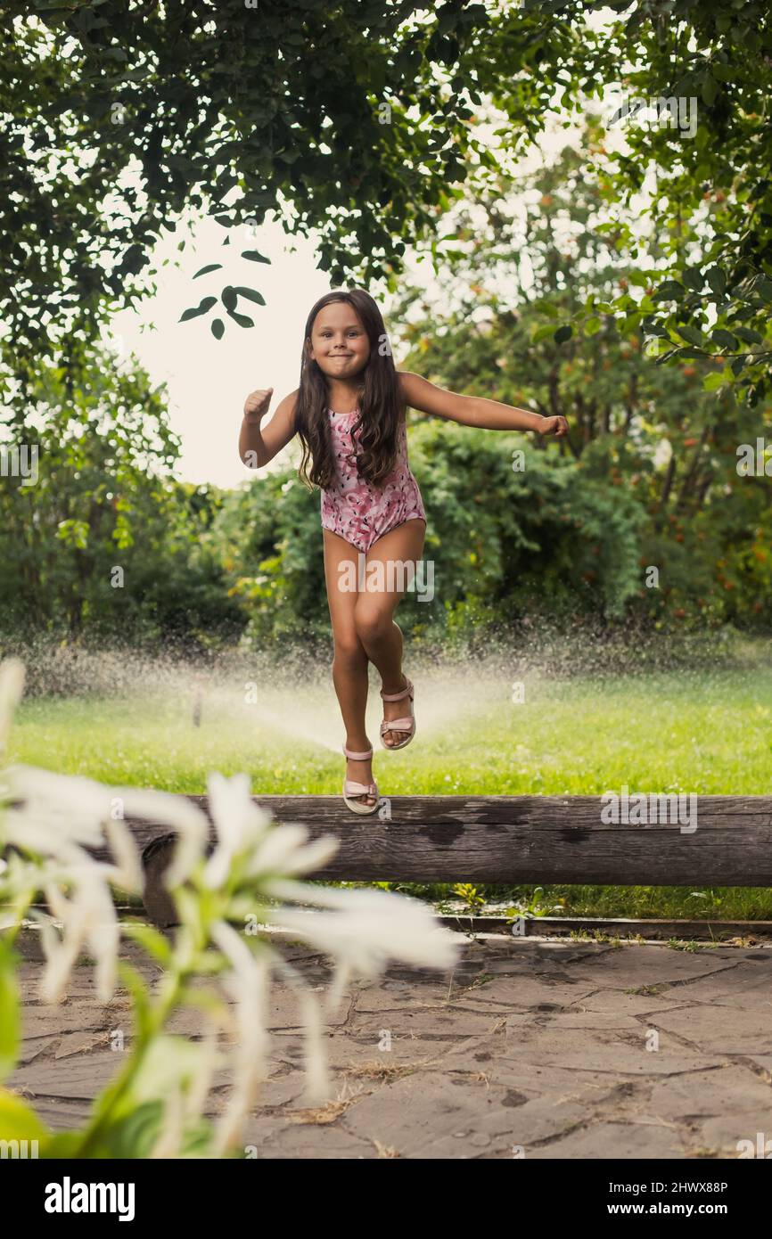 Young shining smiling girl having fun jumping off log, indulging and playing outside in park with trees and bushes in background in daytime. Dreaming Stock Photo