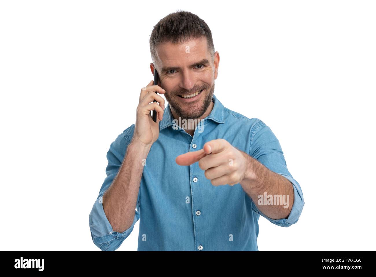 chatty guy in denim shirt having a conversation on the phone, smiling and pointing finger on white background in studio Stock Photo