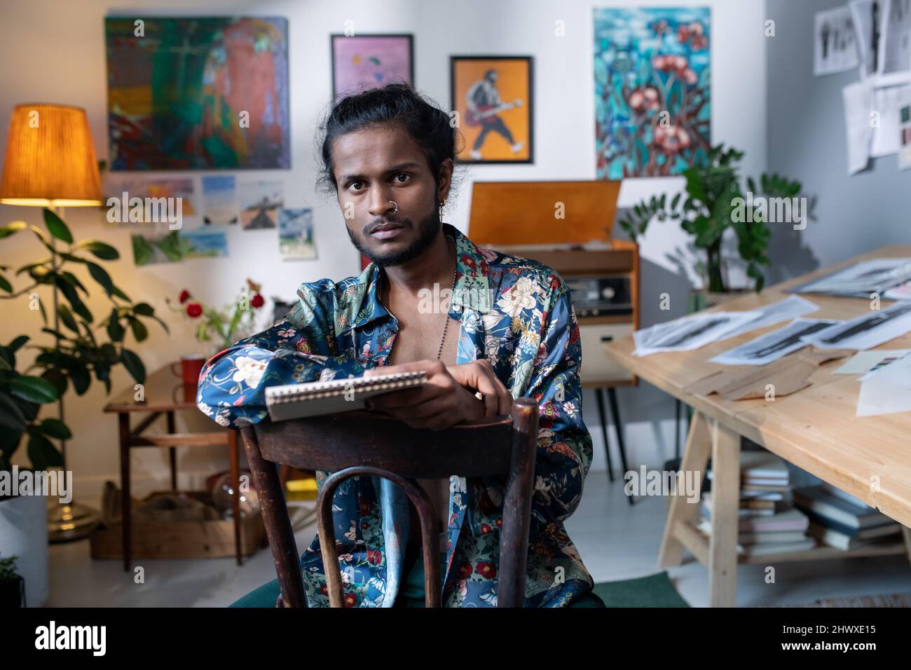 Self-confident young man of Indian ethnicity in smart casualwear working in art studio while sitting against walls with paintings Stock Photo