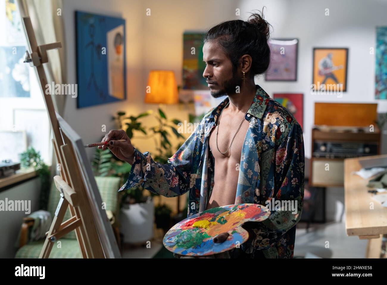 Young contemporary painter with color palette creating new artwork while standing in front of easel in his studio or workshop Stock Photo