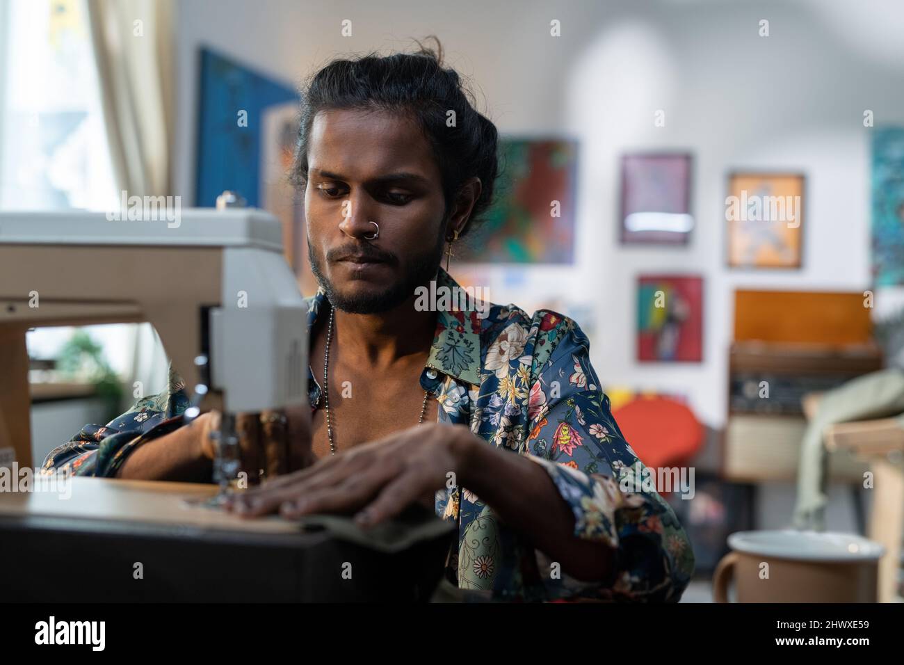 Serious stylish male tailor in non-gendered clothing creating new apparel while sitting in front of camera by electric sewing machine Stock Photo