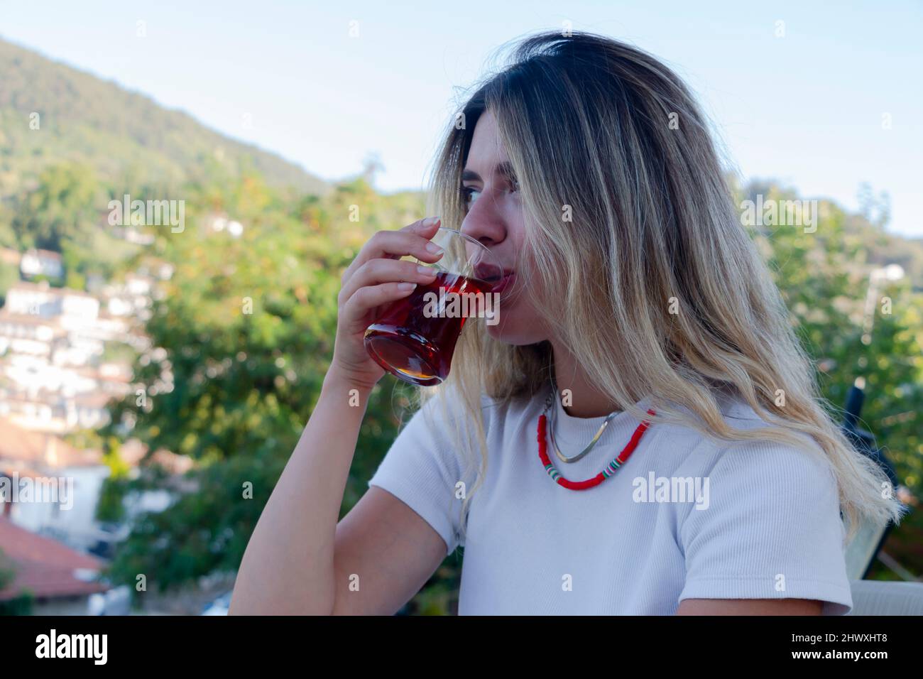 A blonde girl drinking red turkish tea in nature. The weather is sunny. She is looking forward. Stock Photo