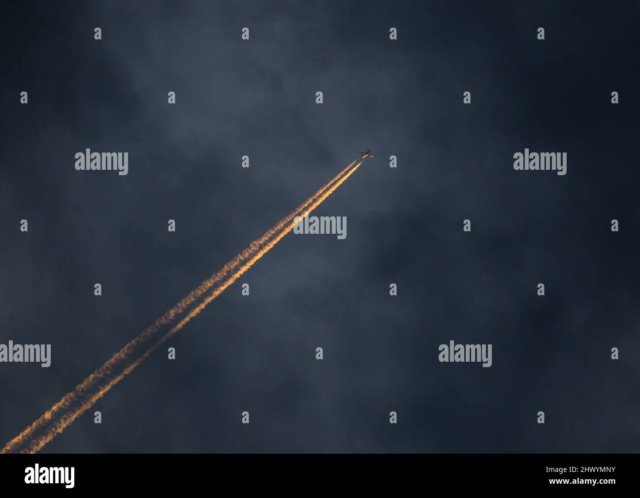 A commercial airliner at high altitude flies at sunset with illuminated contrails. Stock Photo