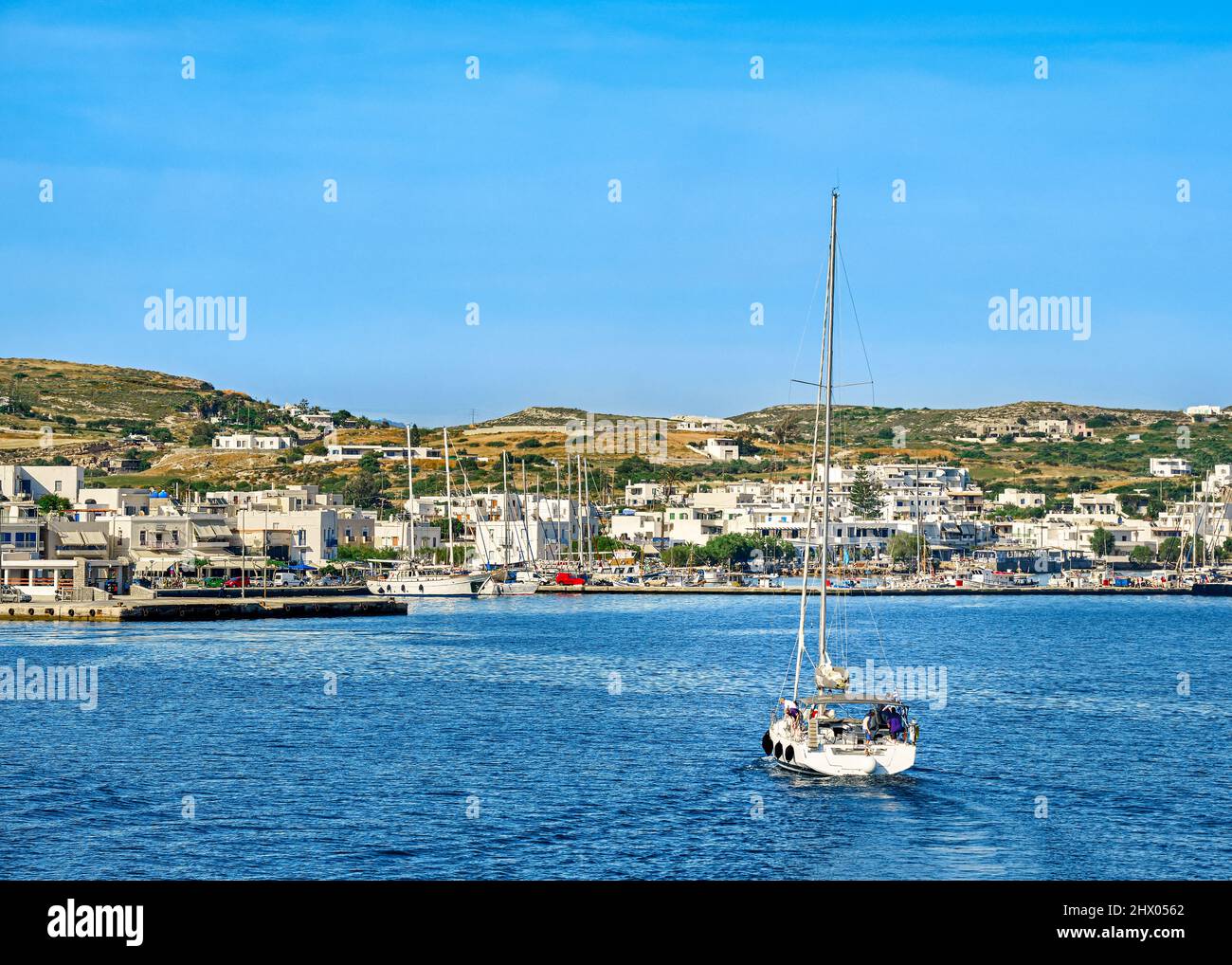 Sailboat going to port of Greek island. Yachts and fishing boats in harbor at sunny summer day. Whitewashed houses on green hills. Stock Photo