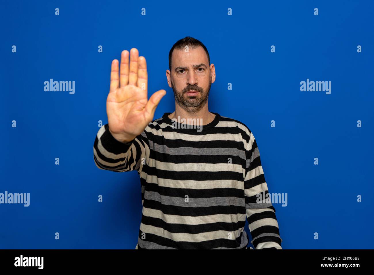 Handsome man wearing striped sweater over isolated blue background stopping singing with palm of hand. Warning expression with negative and serious Stock Photo