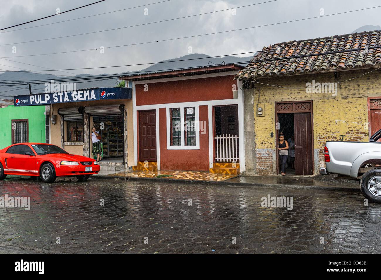 Women shop keepers watch the rain in Jinotega, Nicaragua.  Red Mustang in front of one store. Stock Photo