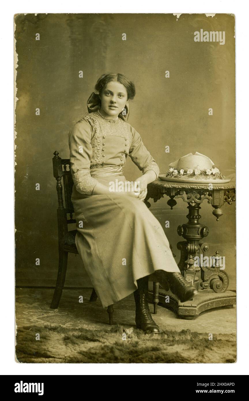 Original WW1 era postcard of pretty teenage girl, wearing a skirt with decorated blouse, and bow in her long hair, with decorated straw hat on the table. From the studio of Barcroft & Crabtree, with studios in Burnley and Nelson, Lancashire, England, U.K. circa 1914. Stock Photo