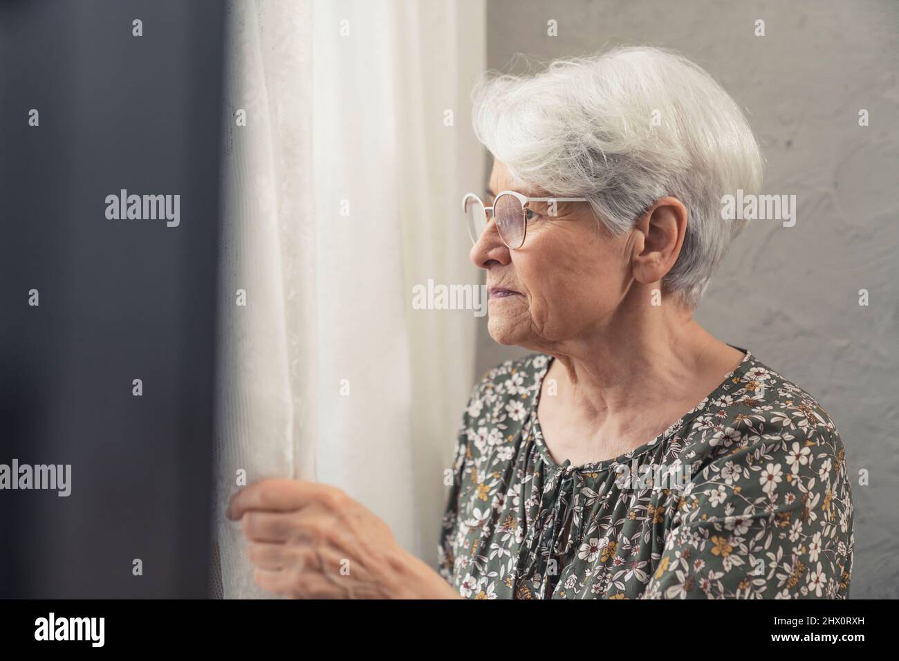 portrait upset caucasian older woman pensioner lady touching curtain gazing through the window. High quality photo Stock Photo