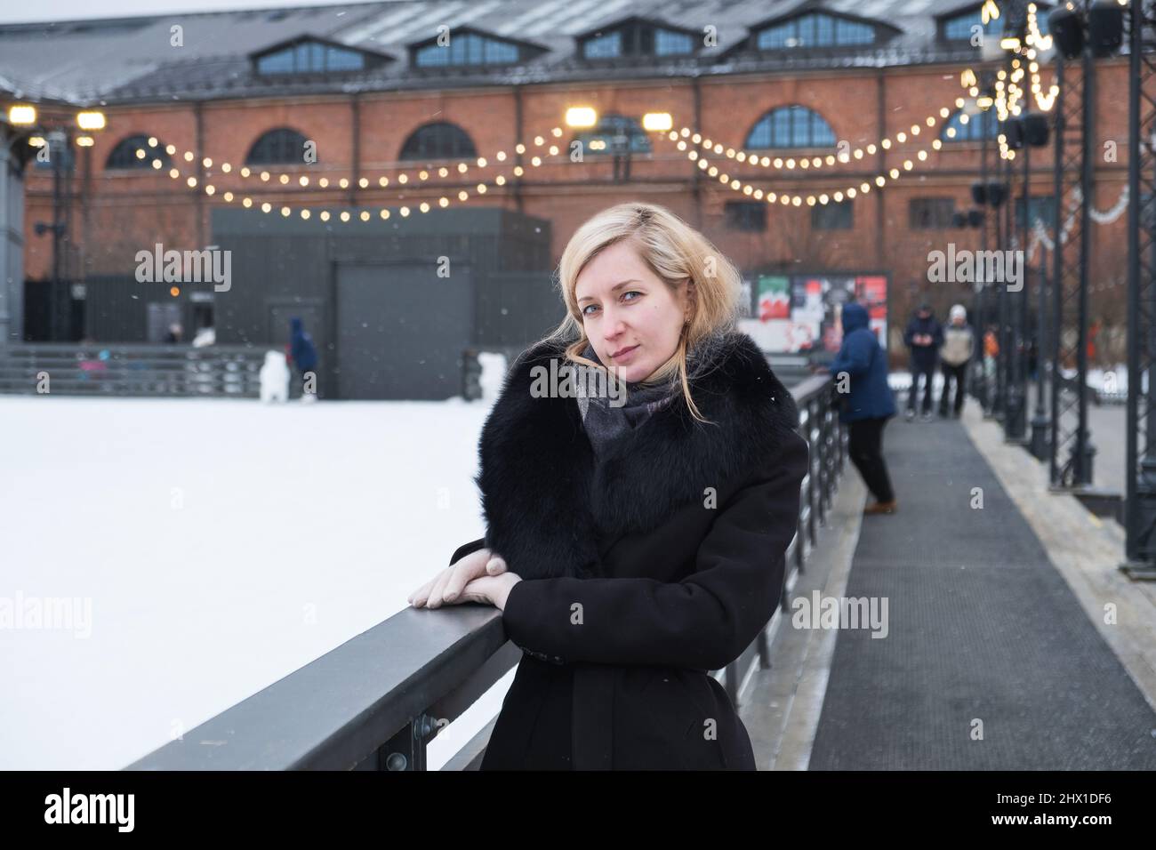 Blonde woman in coat with fur collar stands at side of ice rink. Stock Photo