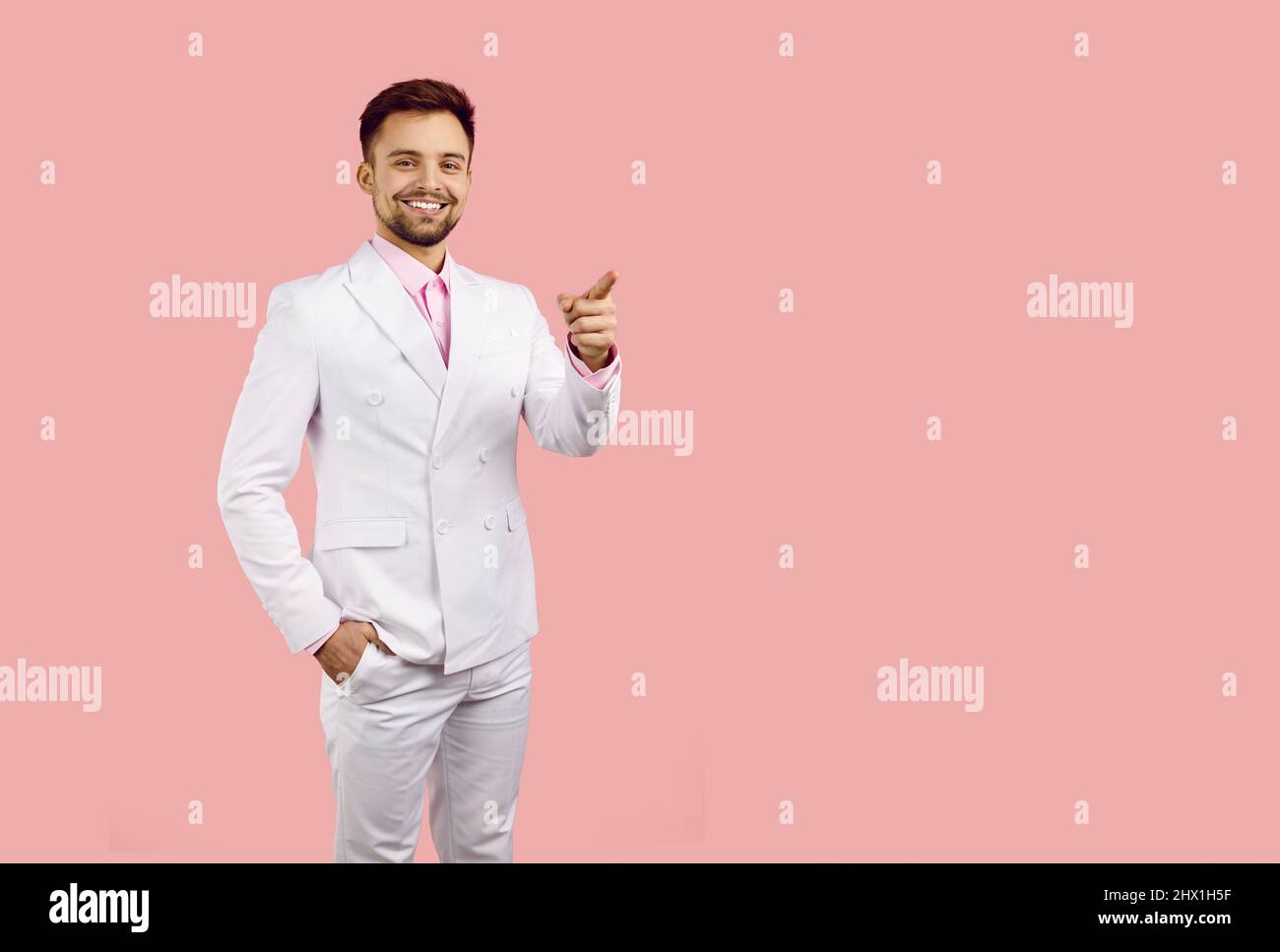 Smiling man in white suit looks at you and points his index finger at copy space on pink background. Stock Photo
